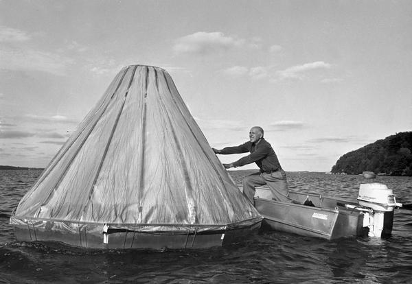 A man sets up a cone-shaped instrument designed to distill fresh water from salt water as he leans out of a small motor boat on the water.