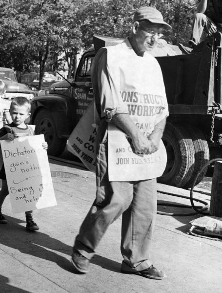 A man wearing a construction workers' union sign pickets in the bright sun, followed by a young boy with a homemade sign of support.