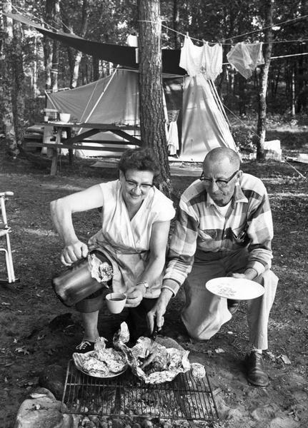 Husband and wife prepare meal on top of a campfire grill.