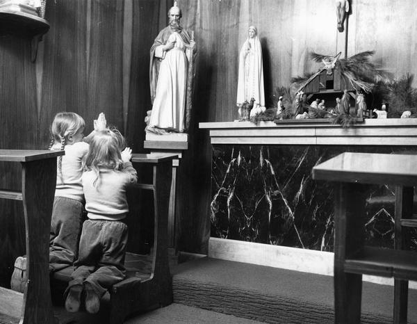 Two little girls kneel before an altar that is holding statues and a creche.