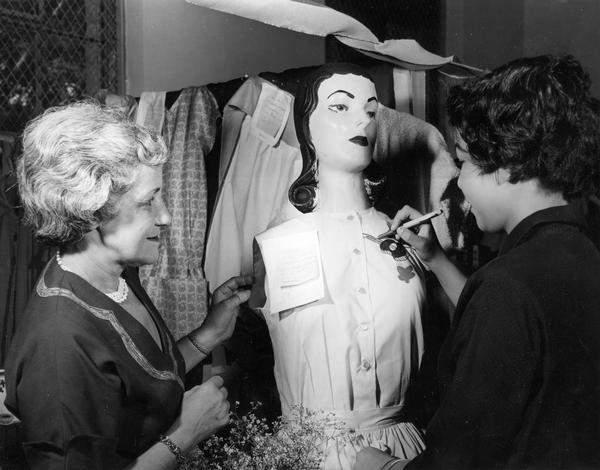 A girl draws a design on a blouse worn by a mannequin while a woman observes her progress.