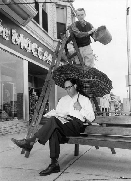 Mounted on a ladder, a man is poised to empty a bucket onto the open umbrella of a man sitting on a sidewalk bench.