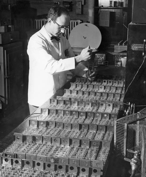 Holding a dropper over racks of test tubes, a man in a lab coat aims to place drops in the receptacles.