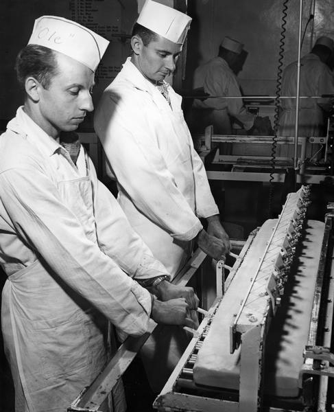 Two men operate a machine that uses wires to make equal portions out of a large slab of butter.