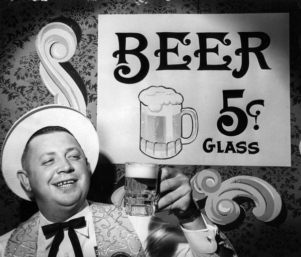 Knights of Columbus member celebrates at an annual "gay nineties" party. He stands next to a sign that advertises beer at five cents a glass.