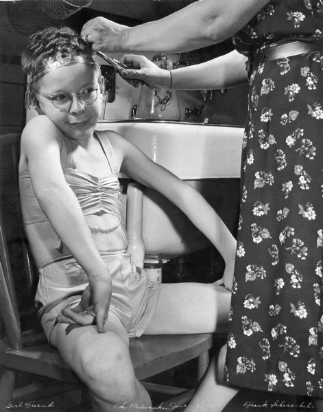 A young boy sits while getting his hair curled with an electric curling iron. According to an article that appeared in the January 9, 1939 <i>Life</i> magazine, he was a member of Boy Scout Troop 1 of Grace Episcopal Church in Sheboygan, which performed two shows of a "burlesque" version of "Romeo and Juliet." All roles were played by boys aged 9-17. The script and songs were written by Father William Elwell, Episcopal minister, who also directed and accompanied on piano. Many liberties were taken with the plot (such as having a happy ending, with Romeo rescuing Juliet from the tomb), and the embellishments of modern slang, songs and dances provided the audience with endless laughs.