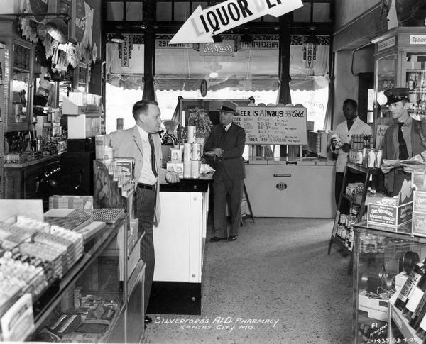 Silverford's A.I.D. Pharmacy, with proprietor, assistant, customer and mailman standing among numerous retail displays and a McCormick-Deering "four in line" beer cooler.