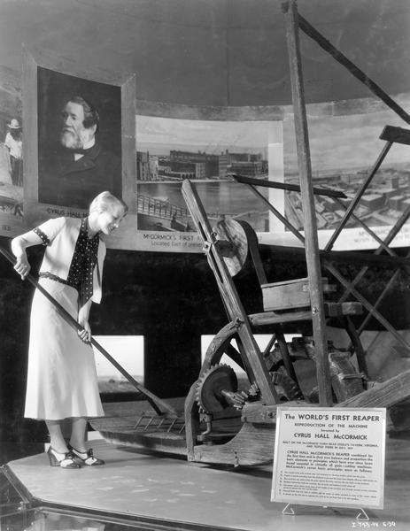 Female model posing with a rake next to a reproduction of Cyrus Hall McCormick's first reaper at International Harvester's "A Century of Progress" Worlds Fair exhibit. A portrait of Cyrus McCormick and photos of his factories are in the background. The reaper replica was most likely produced for the "reaper centennial" in 1931.
