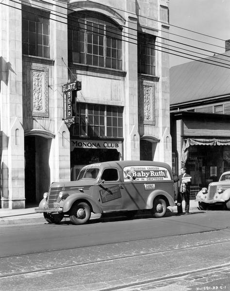 Man unloading boxes of "Baby Ruth" and "Butterfinger" candy bars from an International D-2 truck outside the Monona Club. The truck was owned by the Supreme Specialty Company. The Monona Club building includes a large hanging Blatz beer sign.