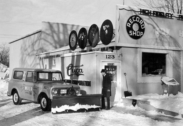 Station manager Ron Nicols and an International Scout truck with plow outside Wisconsin's Very Live Radio station headquarters. WVLR shared quarters with Cuca Records and Sara Sound Studios. James Kirchstein managed the recording company and studio, and was chief engineer and originator of WVLR. Caption reads: "Roving Reporter, Station Manager Nichols leaves WVLR studio, which shares underground quarters with recording company."