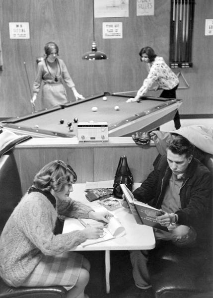 Teenagers studying and playing pool at the Eagle's Nest, a popular teen hangout in downtown Sauk-Prairie. The Eagle's Nest had a juke-box, but the WVLR (Wisconsin's Very Live Radio) radio pictured here was likely to be on, especially during the daily one hour rock 'n roll show. The Petula Clark song "Downtown" was playing when the photograph was taken. Caption reads: "Popular Song, 'Downtown,' accompanies homework session and pool game downtown at the Eagle's Nest, a favorite teenage hangout. Nest has juke-box, but WVLR receiver is more likely to be on, especially during rock-'n-roll show."