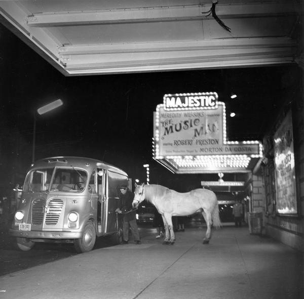 A man hitching Tommy, the Arabian stallion acting horse, to an International Metro AM-120 truck on Broadway as a part of Tommy's stable-to-theatre shuttle. Tommy, part of the Chateau Theatrical Animals Company, was managed by Fred Birkner. A marquee advertising "The Music Man" is in the background.