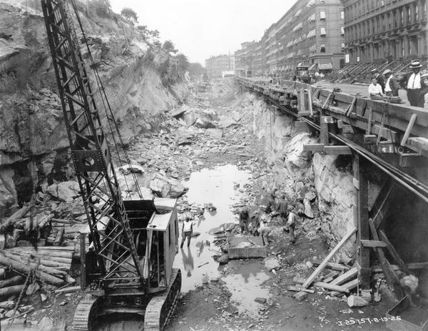 View looking down at workers at work below street level, with cranes and International trucks excavating area for the future New York City subway. On the right people are watching from along a street barrier. Brick buildings are across the street.