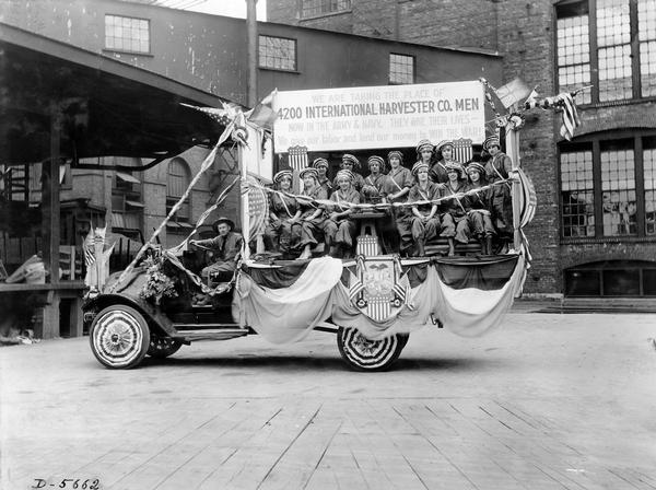 International G-1(?) truck carrying International Harvester Company women workers during World War I. The truck is decorated with flags and shields bearing the stars and stripes. A sign mounted above the women reads: "We are taking the place of 4200 International Harvester Co. men now in the army and navy. They give their lives - we give our labor and lend our money to win the war!"