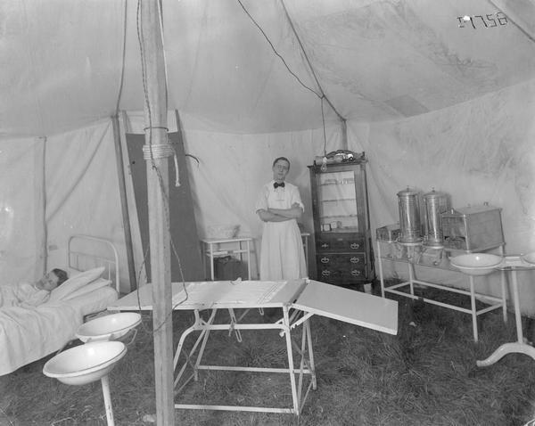 Medical tent with what appears to be a patient in a bed, and doctor standing near a medical cabinet. The photograph is part of an album devoted to International Harvester's McCormick Works. The McCormick Works was built by Cyrus McCormick in 1873 and became part of International Harvester in 1902. The factory was located at Blue Island and Western Avenues in the Chicago subdivision called "Canalport." It was closed in 1961.