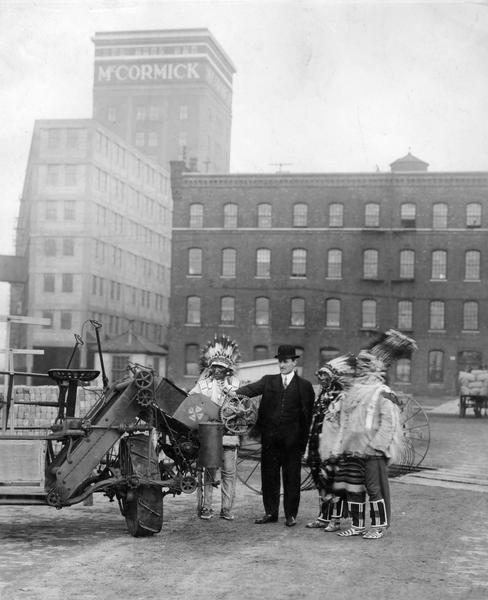Man, most likely R.G. Brooks, showing a Milwaukee grain binder to three Native Americans in traditional dress in front of McCormick Works. One of the Native Americans may be "Chief Lazy Boy."
