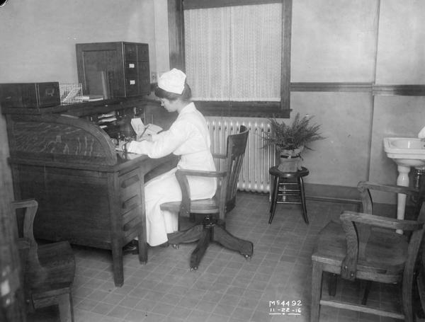 Nurse writing at a desk inside an office at McCormick Works. The McCormick Works was built by Cyrus McCormick in 1873 and became part of International Harvester in 1902. The factory was located at Blue Island and Western Avenues in the Chicago subdivision called "Canalport." It was closed in 1961.