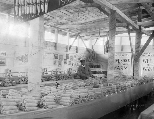 African American man sitting inside an agricultural display of corn, potatoes, canned goods and International Harvester agricultural lecture charts. A banner over the display reads "Tuskegee Institute." Large signs on the wall in the background read: "Live Stock on Every Farm" and "Weeds Mean Waste."