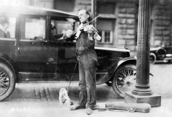 A street musician named Frank Ritter playing violin on a city sidewalk (possibly in Chicago), with a dog leashed to his belt. A tin cup is attached to the violin.