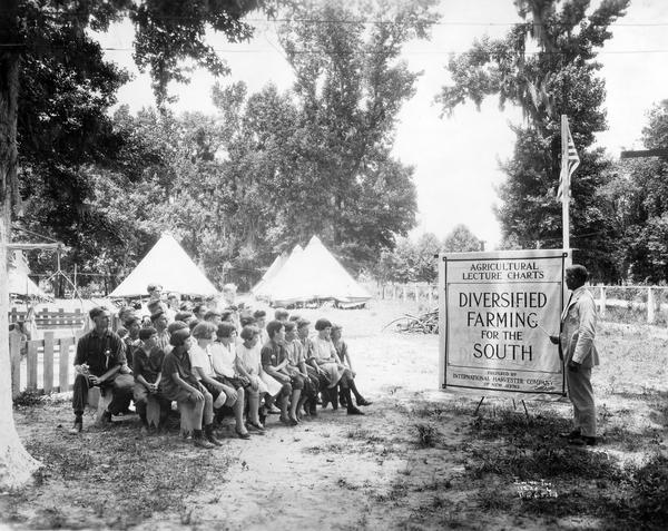 At Louisiana State University, an instructor lectures a group of children on agriculture using charts prepared by International Harvester Company. The lecturer is pointing to a chart with the title "Diversified Farming for the South."