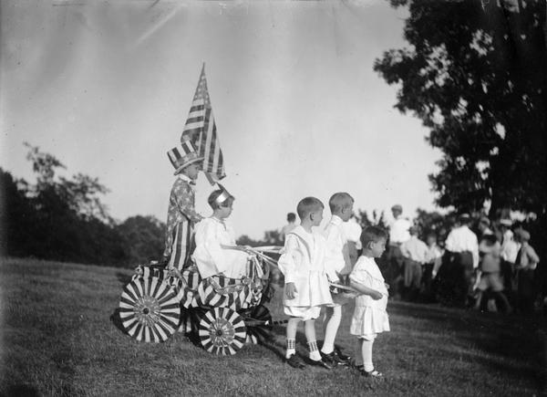 Children dressed up for the Fourth of July at the Trenton Country Club. Three young boys are pulling a girl and boy in a wagon adorned with stars and stripes.