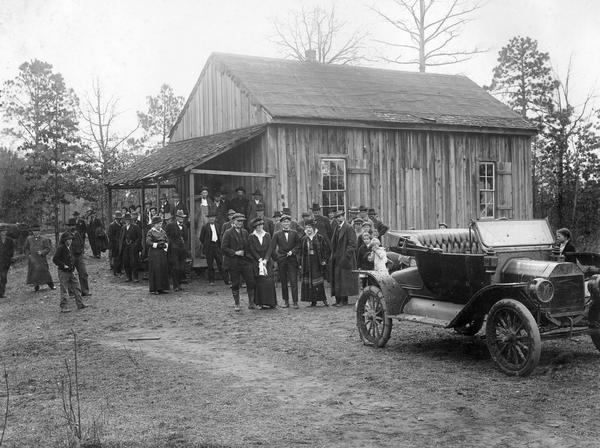 Townspeople and a Ford automobile in front of a one-room schoolhouse in rural Alabama. The original caption reads: "This school house at Alpine, Ala., is a fair sample of the condition in which we found most country schools. It consists of one room, furnished with home made desks. There are no out buildings for the children. Drinking water is furnished from a wooden bucket with a dipper in it for the use of all."