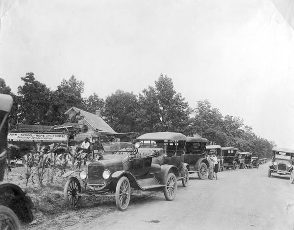 Automobiles, including a Ford runabout, parked along a rural dirt road for an International Harvester Agricultural Extension Department short course and machine demonstration. A sign mounted on a manure spreader reads: "Farm - School - Home Short Course - Machine Demonstration." Two barefoot boys are leaning against one of the cars.