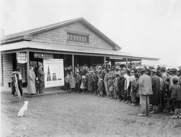 Lindsey Reese of International Harvester's Agricultural Extension Department delivering a lecture on livestock and agriculture to a crowd of people and a dog outside a rural post office and general store.