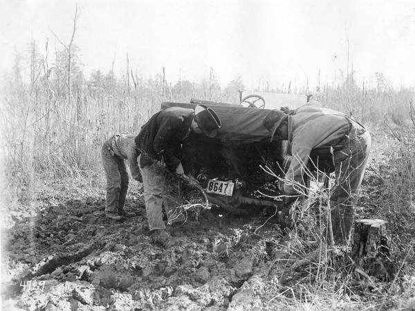 Three employees of International Harvester's Agricultural Extension Department working to free a car stuck in a muddy rural road. The original caption reads: "Many times the roads were not of the best and the speakers were greatly delayed in making their appointments. In going from Turnpike school to Springville school near Pontotoc, Mississippi just such roads were encountered but after an hour's hard work our difficulties were overcome and we were able to make our next engagement notwithstanding the mud on our clothes."
