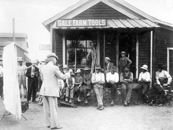 International Harvester Agricultural Extension Department employee W.R. Baughman talking to a small meeting in Dupage County during the Alfalfa Campaign. A group of men and boys are gathered in front of a building that has a sign that reads: "Gale Farm Tools". The company's policy, developed by Professor Holden, was to go to the country to hold meetings and lectures. The policy is described as follows in the original caption: "It is better to talk to a small crowd of fifteen interested farmers that really want information than to have a crowd of two or three thousand that don't care anything about growing alfalfa, or that are not interested in the proposition. It is the Holden Ideas brought in again in the play <i>[sic]</i> of going through the country to hold their meetings. If you want to help people go where they are to help them with their own problems under their own conditions."