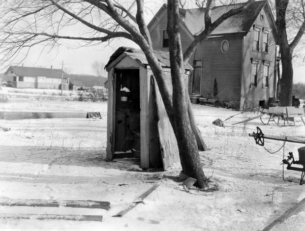 An outhouse without a door during the winter. The outhouse is situated under a tree behind a farmhouse.