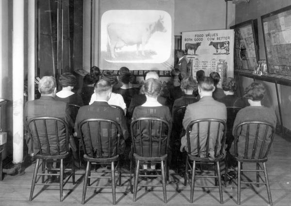 Rear view of men and women sitting in wooden chairs viewing a lantern slide projection of a cow during a presentation by a lecturer from International Harvester's Agricultural Extension Department.