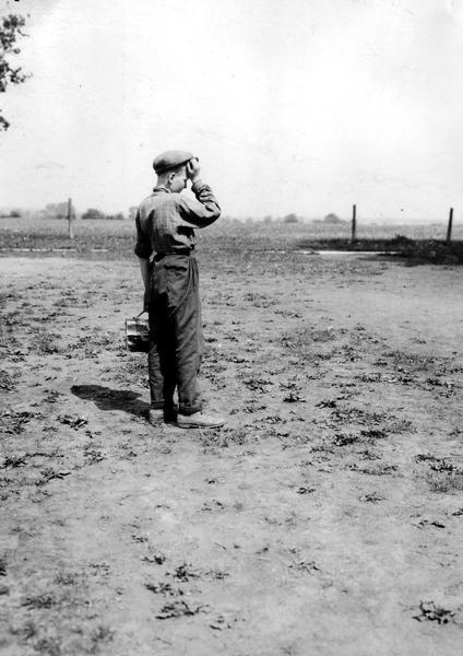 A young boy holding a lunch box has his hand on his head while standing on the grounds of Viall School.