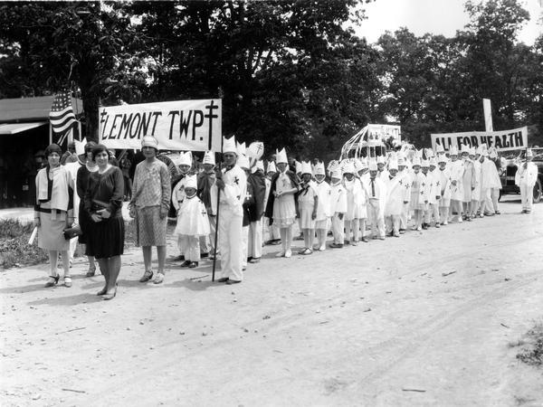 School children and teachers from Lemont township marching in a rural school festival parade. The children are dressed in white cloaks and hats bearing a medical symbol and are carrying a sign with the words "All For Health".