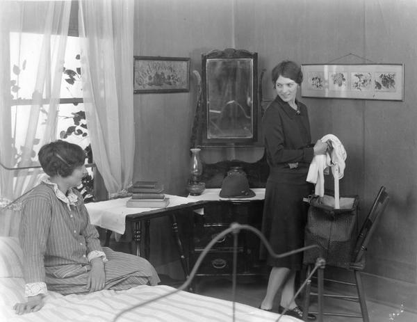 Two young women talking in a bedroom. One is packing or unpacking clothes into a travel bag. A copy of Herbert Quick's "Vandemark's Folly" is among the books on an end table.