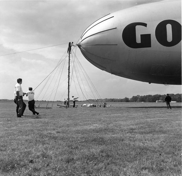 Goodyear "America" blimp lands after final ground check in a field near Wingfoot Lake. The blimp is tied to portable landing mast with generator, machine shop and helium purification unit standing by.