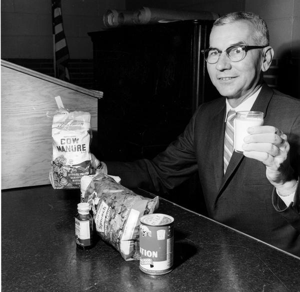 Milton C. Geuther, Illinois American Dairy Association Manager, holding a glass of milk in one hand and a bag of cow manure in the other. Geuther claimed that milk was the best value for the American household. He claimed that each item shown in the photograph was more expensive by the pound than milk.