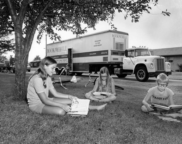 Three children reading books near a bookmobile parked in a suburban neighborhood. The bookmobile was an International Loadstar 1600.