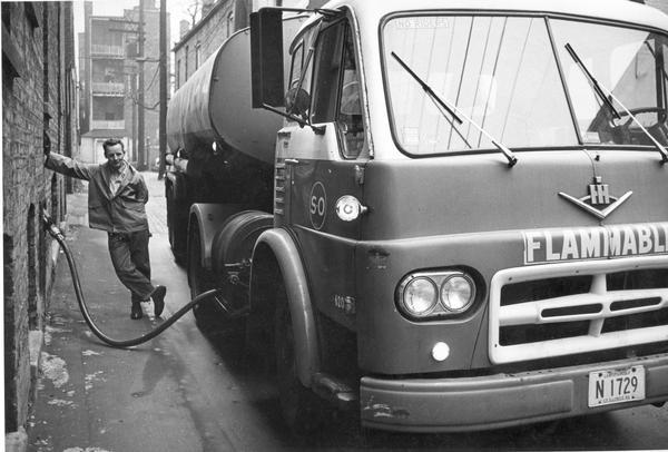 A deliveryman relaxes while fuel oil flows from an International 620 fuel truck to an urban residential home.