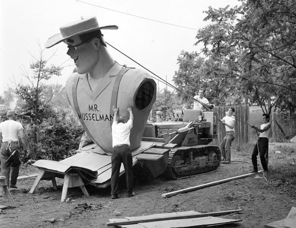 Five men preparing to lift a bust of "Mr. Musselman" with an International TD-6 crawler tractor and Drott high-lift bucket loader. The "Mr. Musselman" bust was part of the Fantasyland Park exhibit at the centennial celebration of Lincoln's Gettysburg address. When completed, Mr. Musselmann stood 22 feet tall, publicizing the apple products of the Musselman Division of Pet Milk Company.