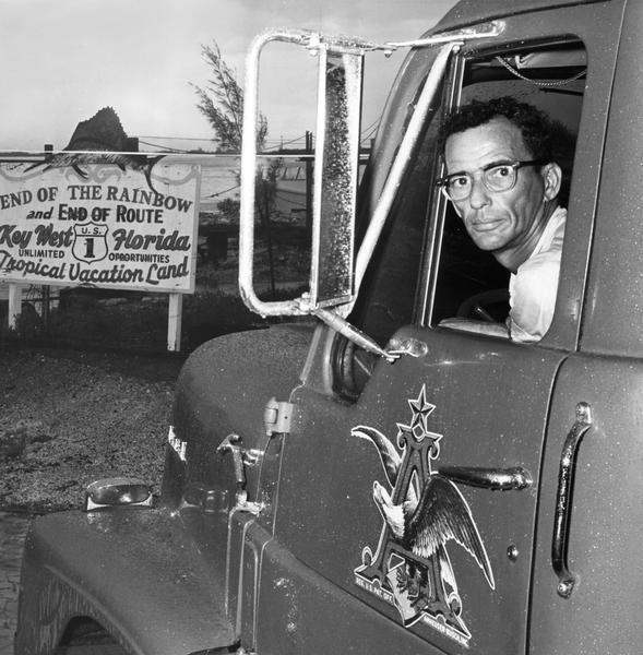 Truck driver Joseph Gonzalez in his International DB-405 Budweiser beer truck at the terminus of U.S. Highway 1. A sign in front of the truck reads "end of the rainbow and end of route U.S. 1; Key West, Florida; unlimited opportunities; tropical vacation land."