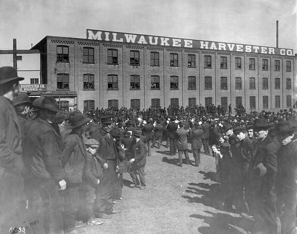 A large crowd assembled outside International Harvester's Milwaukee Works. The crowd is composed mostly of men, but includes a few boys and at least one girl. They appear to be listening to a brass band. The factory was owned by the Milwaukee Harvester Company until 1902.
