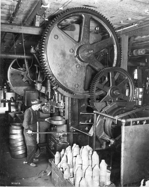A worker at International Harvester's Milwaukee Works draining fluid from a machine with a large gear poised over his head. The factory was owned by the Milwaukee Harvester Company until 1902.