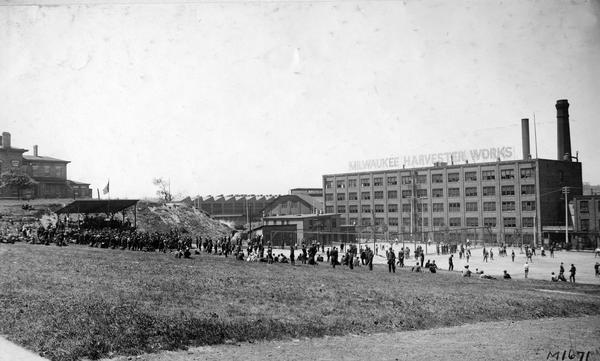 Workers relaxing outside International Harvester's Milwaukee Works. One group is crowded around a stage listening to a band while others are playing baseball, tennis or sitting on the grass. The original caption said: "band playing and games in progress; noon scene on Milwaukee Works playground." The factory was owned and operated by the Milwaukee Harvester Company until 1902.
