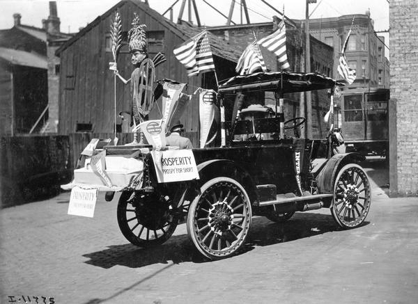 International Model E highwheeler truck adorned with American flags, advertising banners, and a statue of a sprite named "Prospy." "Prospy" was a little man with a corn cob body and a shield shaped like the International Harvester logo. Signs on the truck read: "Prosperity, Prospy for short." The Model E was introduced in the spring of 1915.  Rated at 800 pounds carrying capacity, the E featured an all-steel frame and side flange detachable solid tires. The E, produced until 1916, was priced at $950.