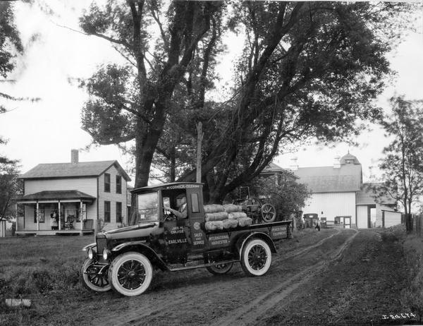 International "Red Baby" (model S) truck loaded with binder twine(?) and a stationary engine leaving a rural farmstead. In the background a woman holding a small child stands watching from the front porch of her house. The truck was owned by McCormick-Deering dealership of Joseph H. Cruise.