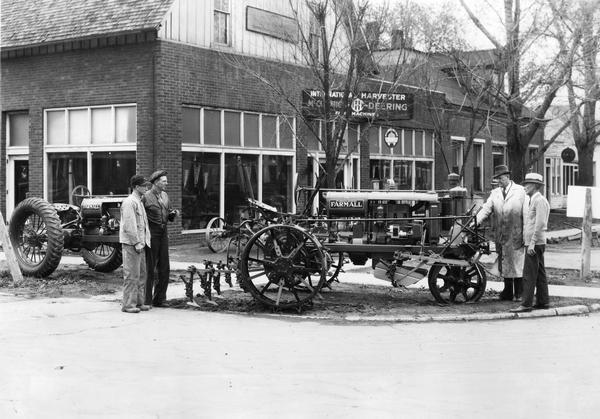 Farmall F-20 tractor with steel wheels parked outside a dealership owned by Gary T. Vogelaar.  At the front of the F-20 Mr. Vogelaar is pointing out to Simon De Haan the automatic shift on the cultivator.  At the rear of the tractor Gary's brother Ben is showing Ray De Haan the new cultivator gangs. A Farmall F-12 tractor with rubber wheels sits in the background.