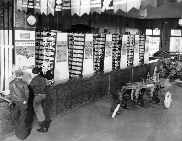 Two customers standing at the parts counter of a McCormick-Deering dealership owned by C. Scheler and Son. Two men are behind the counter. To the right of the counter is a McCormick-Deering disk plow.