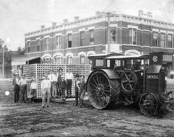Ten men and boys are posing behind a Mogul 15-30 tractor in front of the Bank of Kiowa and the H.G. Waltner Mercantile Company. The tractor is hitched to a large rectangular pen mounted on a skid made of timbers.