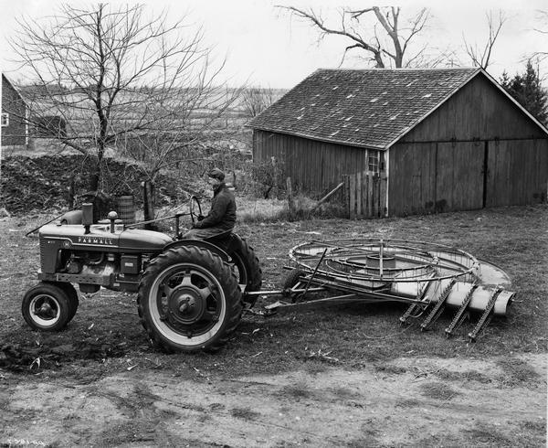 Slightly elevated view of farmer Axel Anderson in his barnyard on a Farmall H tractor with a hemp turning machine. The hemp turning machine was made by the Tower Co. of Mendota, Illinois. The machine picks up the hemp and elevates it to a turntable where it is taken to the other side and dropped back to the ground, turning as it falls.
